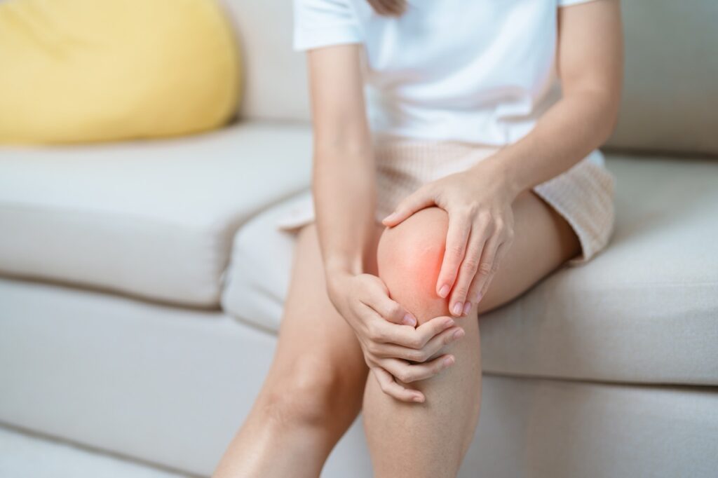 A woman sitting on a white couch at home, holding her painful left knee, highlighted in red, with both hands.