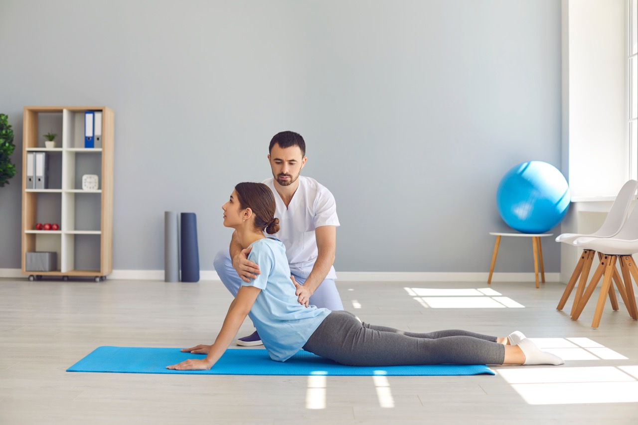 A young female athlete on a blue mat doing back-bending exercise with male physical therapist after a sports injury.